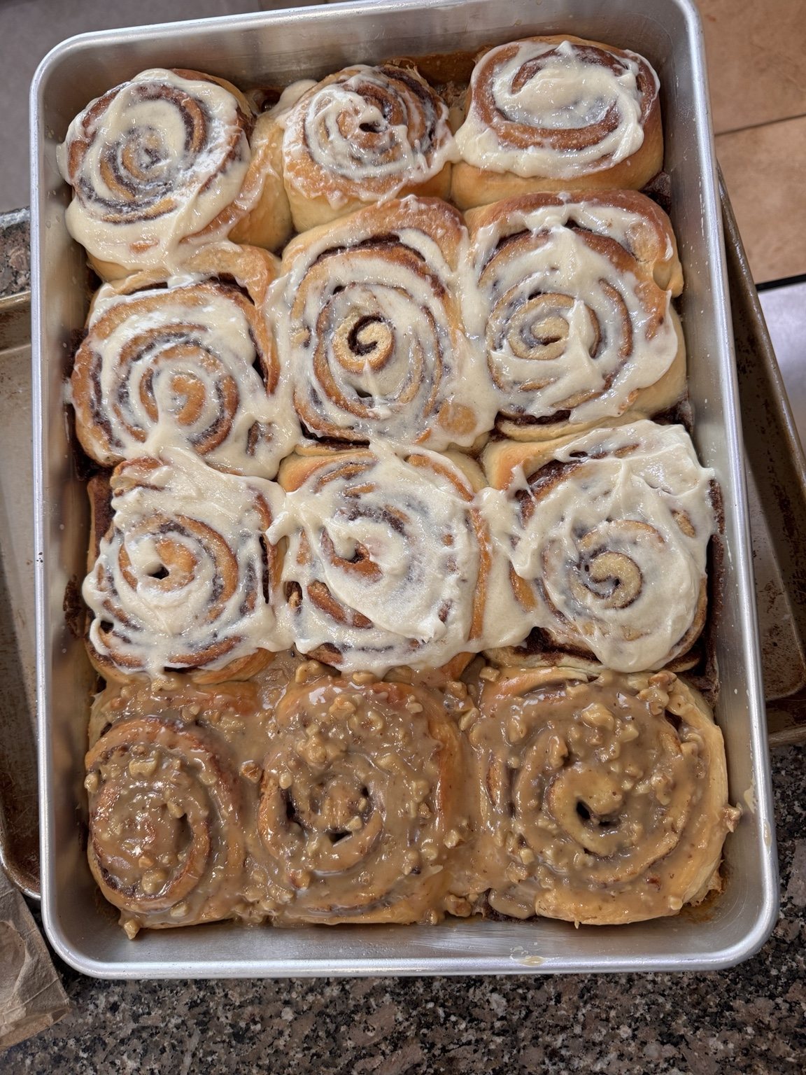 A pan of finished giant cinnamon rolls, most topped with white icing and a few along the bottom with walnut glaze.
