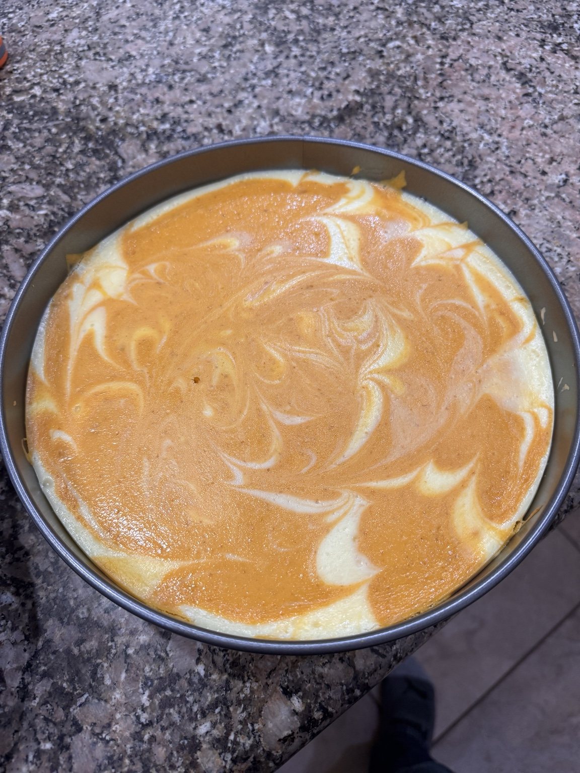 A baked pumpkin swirl cheesecake in a round springform pan, with orange pumpkin and vanilla filling marbled across the top.